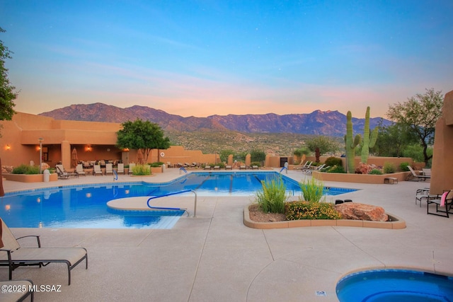 pool at dusk with a hot tub, a mountain view, and a patio