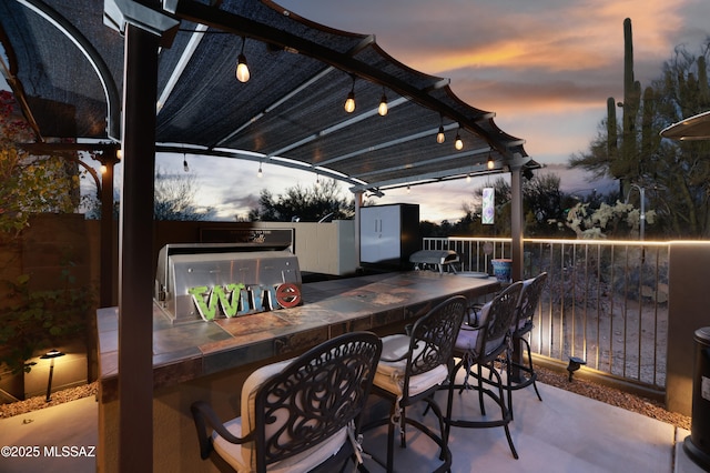 patio terrace at dusk featuring exterior kitchen, a pergola, and a bar