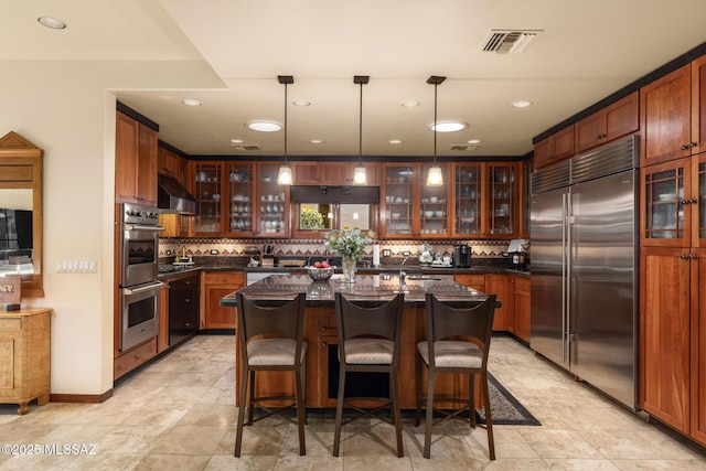 kitchen featuring a kitchen bar, hanging light fixtures, an island with sink, stainless steel appliances, and wall chimney range hood