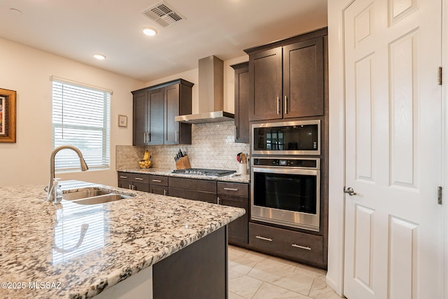 kitchen with sink, dark brown cabinetry, light stone counters, stainless steel appliances, and wall chimney exhaust hood