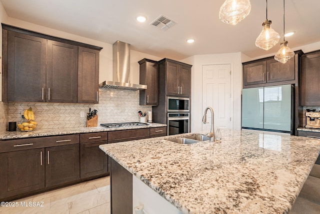 kitchen with appliances with stainless steel finishes, sink, light stone counters, a center island with sink, and wall chimney exhaust hood