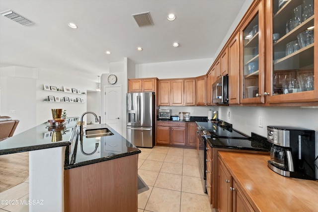 kitchen featuring sink, light tile patterned floors, appliances with stainless steel finishes, dark stone countertops, and a center island with sink