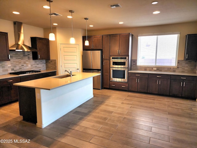 kitchen featuring wall chimney exhaust hood, sink, dark brown cabinets, a center island with sink, and stainless steel appliances