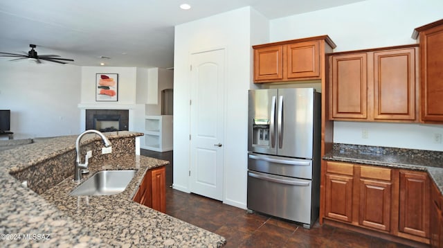 kitchen featuring sink, stainless steel fridge, a tile fireplace, ceiling fan, and dark stone counters