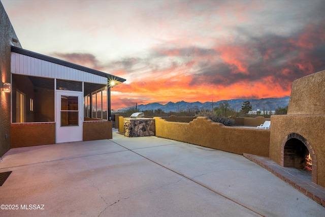 patio terrace at dusk featuring a mountain view, a sunroom, an outdoor fireplace, and exterior kitchen