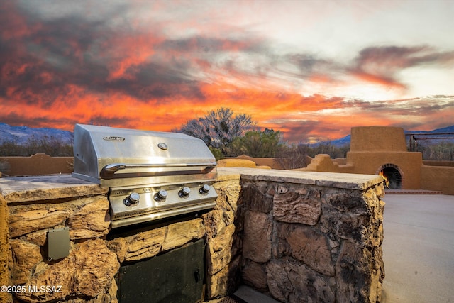 patio terrace at dusk featuring an outdoor kitchen, grilling area, a mountain view, and an outdoor stone fireplace