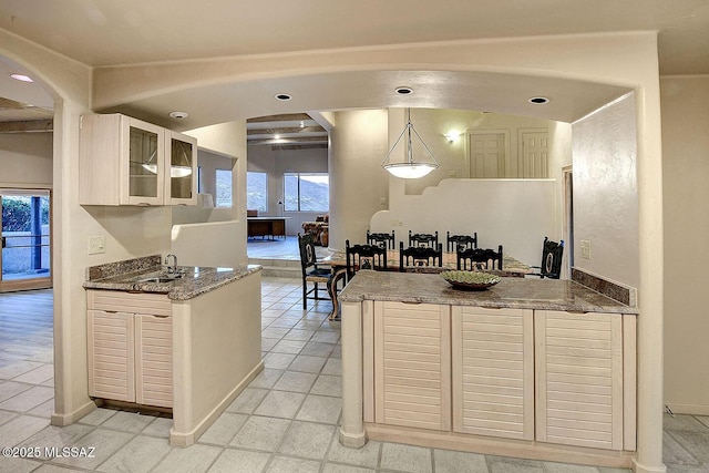 kitchen featuring arched walkways, glass insert cabinets, decorative light fixtures, stone counters, and a sink