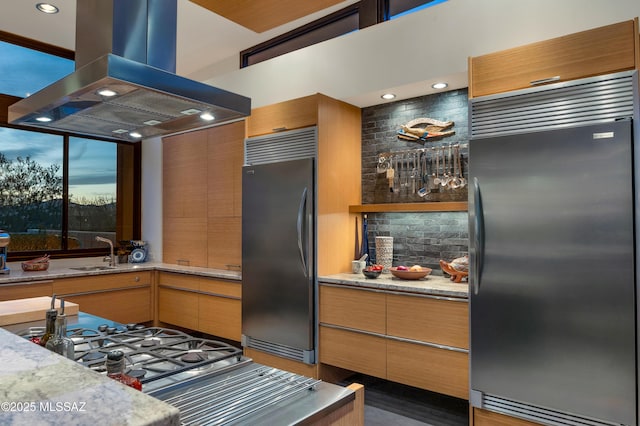 kitchen with a sink, built in refrigerator, island exhaust hood, and light stone countertops