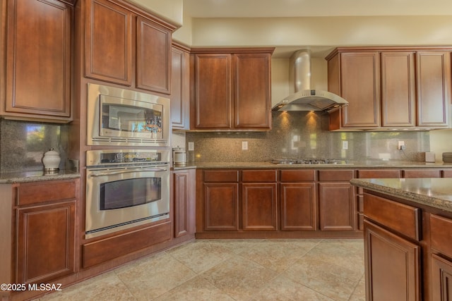 kitchen featuring wall chimney exhaust hood, appliances with stainless steel finishes, decorative backsplash, and dark stone counters