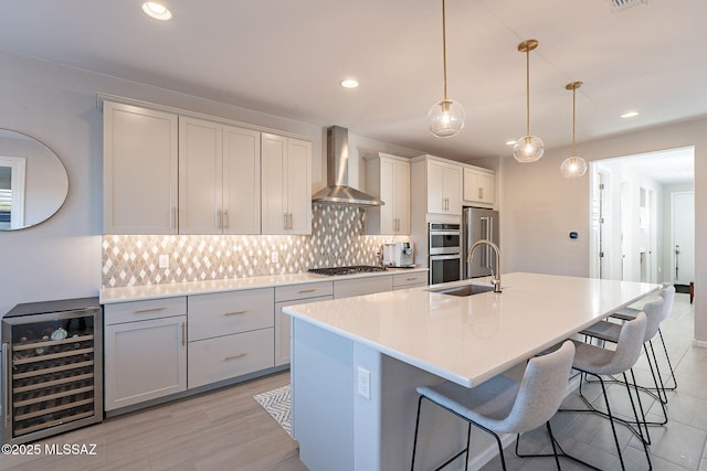 kitchen with wine cooler, tasteful backsplash, a center island with sink, pendant lighting, and wall chimney range hood