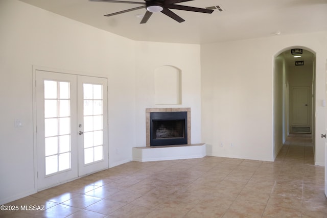 unfurnished living room featuring ceiling fan, arched walkways, a tiled fireplace, and french doors