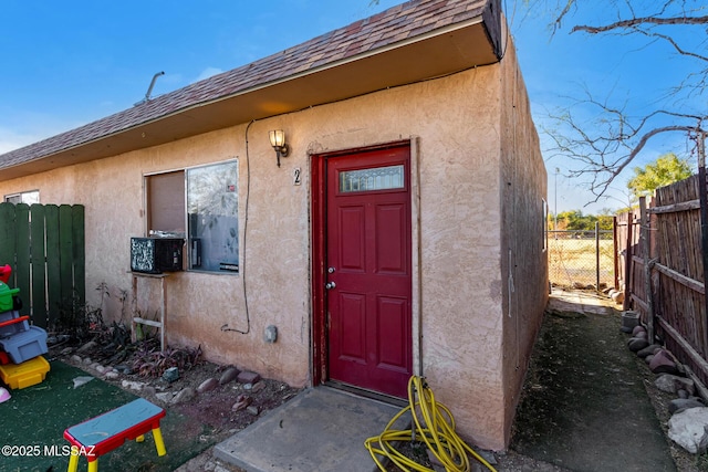 doorway to property featuring fence, roof with shingles, and stucco siding