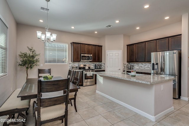 kitchen featuring appliances with stainless steel finishes, decorative light fixtures, a kitchen island with sink, light tile patterned floors, and light stone countertops