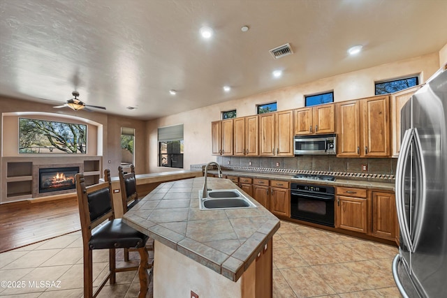 kitchen with sink, a kitchen island with sink, stainless steel appliances, light tile patterned flooring, and tile countertops