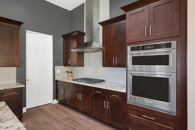 kitchen with appliances with stainless steel finishes, hardwood / wood-style floors, light stone counters, decorative backsplash, and wall chimney exhaust hood