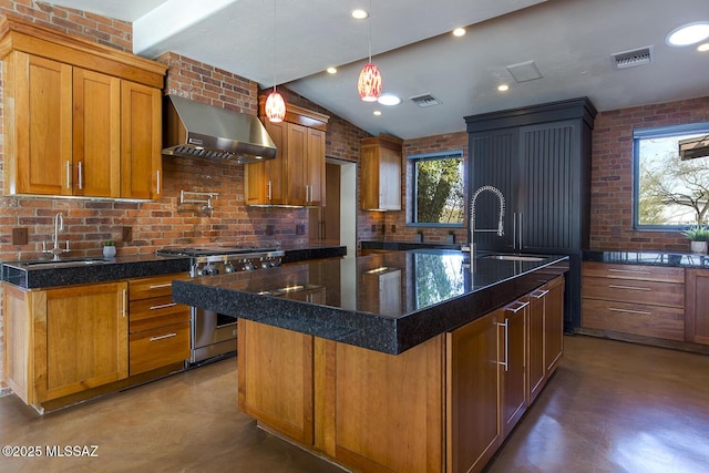 kitchen featuring stainless steel stove, a kitchen island with sink, a sink, wall chimney exhaust hood, and brown cabinetry