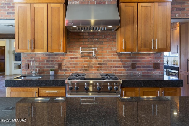 kitchen featuring stove, brown cabinetry, a sink, dark stone counters, and wall chimney exhaust hood