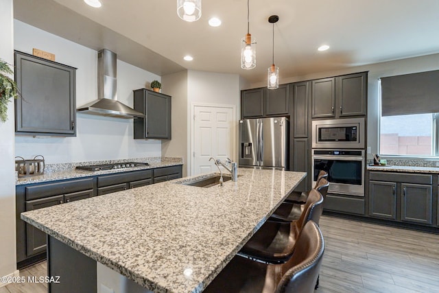 kitchen with appliances with stainless steel finishes, pendant lighting, an island with sink, wall chimney range hood, and light wood-type flooring