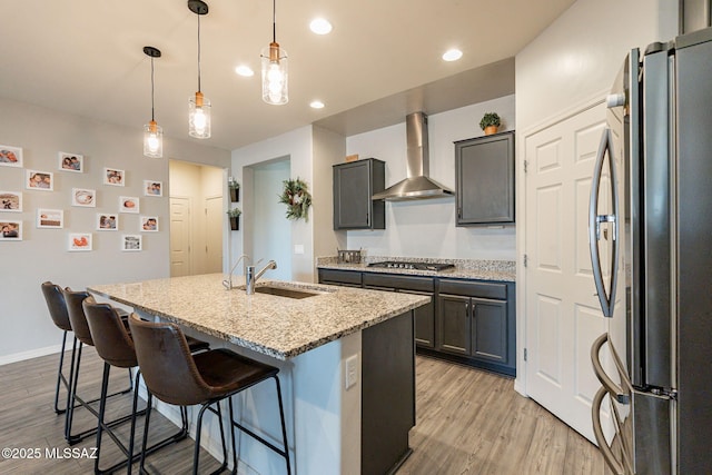 kitchen with sink, an island with sink, pendant lighting, stainless steel appliances, and wall chimney range hood