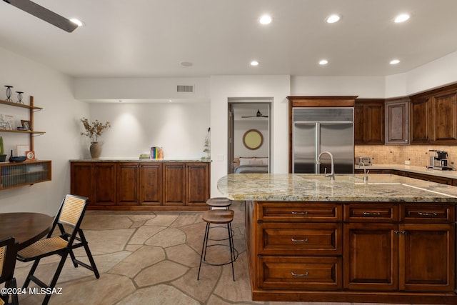 kitchen featuring built in fridge, a kitchen breakfast bar, decorative backsplash, light stone countertops, and a center island with sink