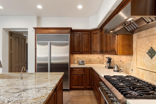 kitchen featuring wall chimney range hood, sink, high end appliances, light stone counters, and tasteful backsplash