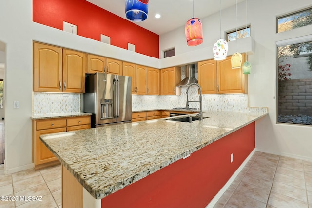 kitchen featuring hanging light fixtures, kitchen peninsula, stainless steel refrigerator with ice dispenser, light stone countertops, and wall chimney range hood