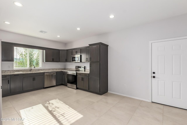 kitchen with appliances with stainless steel finishes, sink, light tile patterned floors, light stone counters, and dark brown cabinets