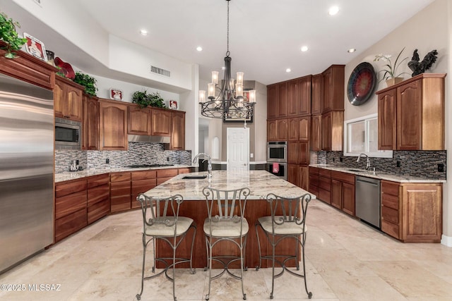 kitchen with sink, light stone counters, built in appliances, decorative light fixtures, and a center island with sink