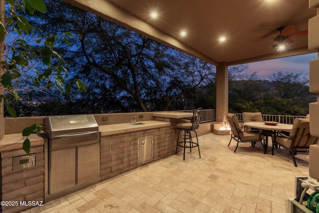 patio terrace at dusk featuring grilling area, an outdoor kitchen, sink, and ceiling fan