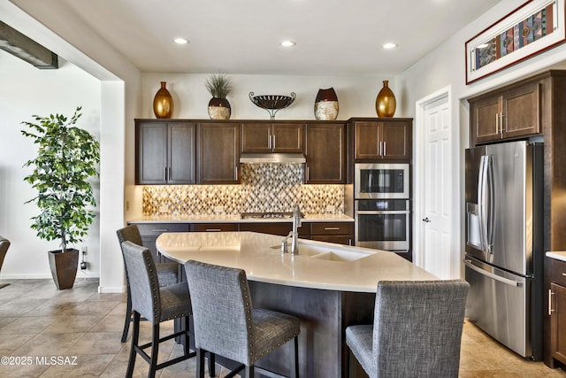 kitchen with appliances with stainless steel finishes, tasteful backsplash, an island with sink, sink, and dark brown cabinetry