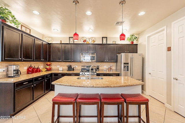 kitchen with sink, appliances with stainless steel finishes, hanging light fixtures, light stone counters, and an island with sink