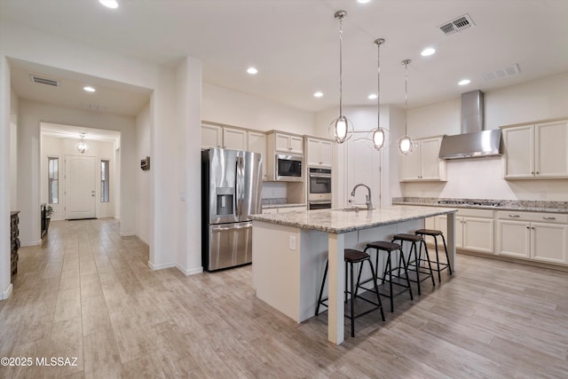 kitchen featuring appliances with stainless steel finishes, pendant lighting, an island with sink, light stone counters, and wall chimney exhaust hood