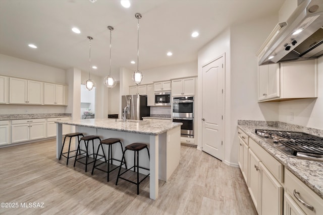 kitchen with an island with sink, wall chimney exhaust hood, light wood-style flooring, appliances with stainless steel finishes, and a breakfast bar area