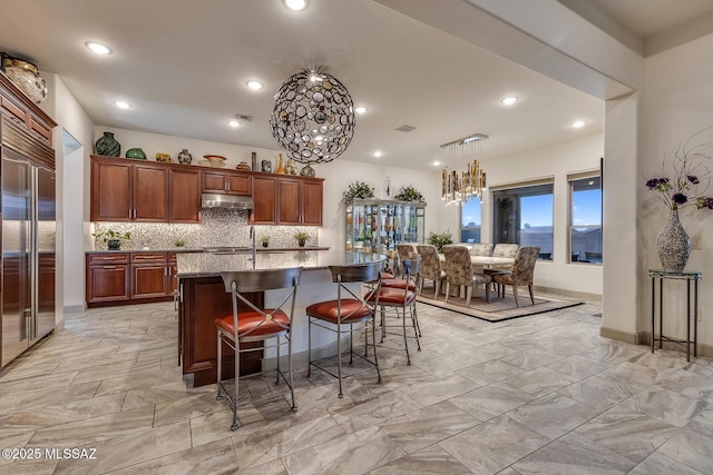 kitchen featuring a breakfast bar, hanging light fixtures, a kitchen island with sink, high quality fridge, and under cabinet range hood