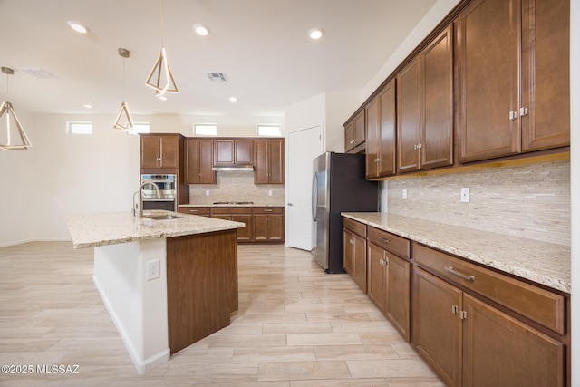 kitchen featuring appliances with stainless steel finishes, decorative light fixtures, sink, light stone counters, and a center island with sink