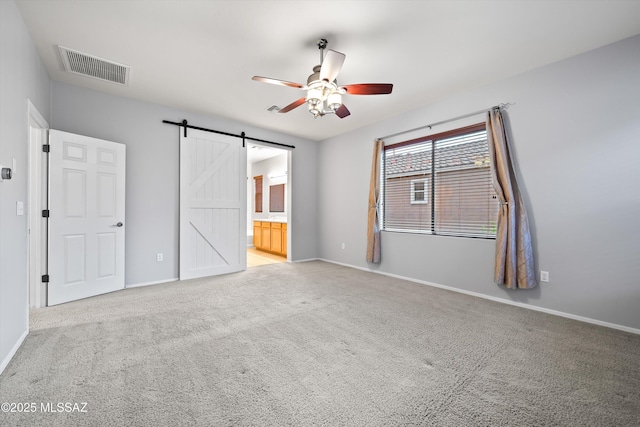 unfurnished bedroom featuring ceiling fan, light colored carpet, a barn door, and ensuite bath