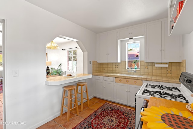 kitchen featuring white range with gas stovetop, white cabinetry, tile countertops, light tile patterned floors, and decorative backsplash