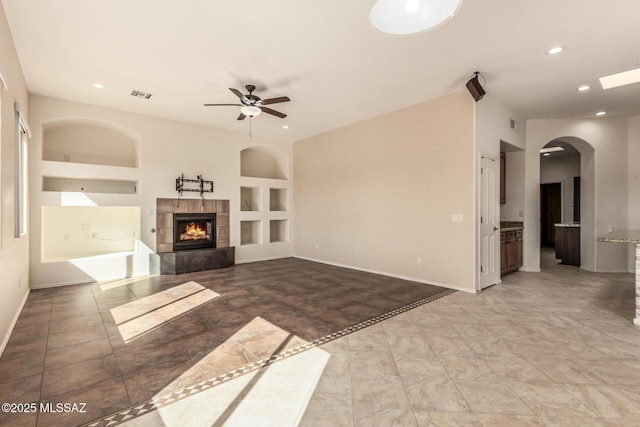 unfurnished living room with a tiled fireplace, built in features, and ceiling fan