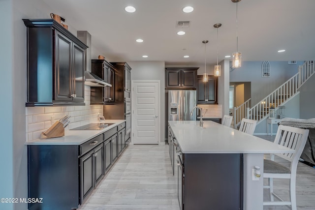 kitchen with a kitchen island with sink, hanging light fixtures, stainless steel appliances, a kitchen bar, and wall chimney exhaust hood