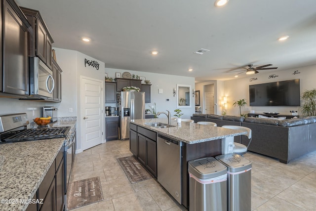 kitchen featuring sink, a kitchen island with sink, dark brown cabinetry, stainless steel appliances, and light stone countertops