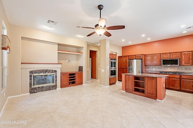 kitchen with appliances with stainless steel finishes, a fireplace, decorative backsplash, ceiling fan, and a center island with sink