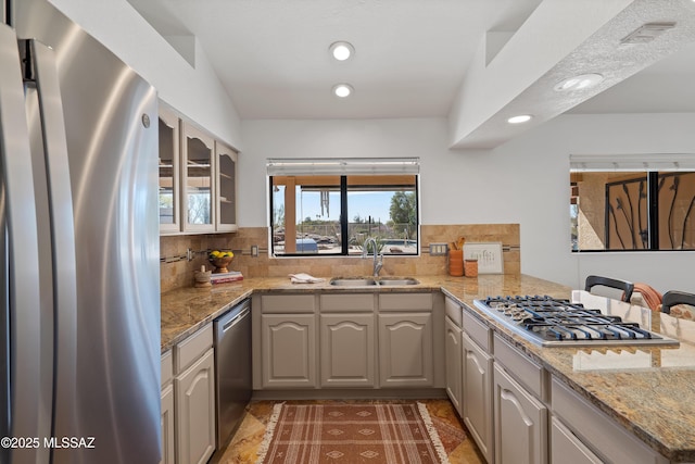 kitchen with light stone counters, appliances with stainless steel finishes, glass insert cabinets, a sink, and a peninsula