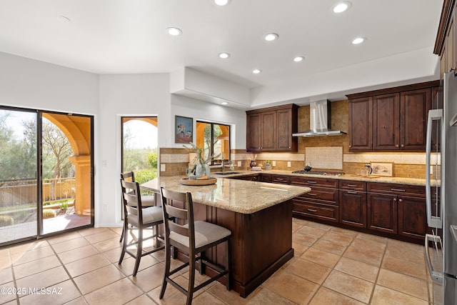 kitchen featuring a breakfast bar area, kitchen peninsula, stainless steel appliances, light stone countertops, and wall chimney range hood