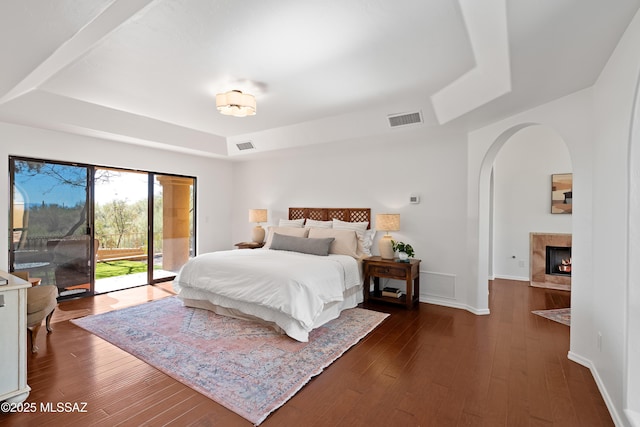 bedroom featuring a tiled fireplace, a tray ceiling, dark hardwood / wood-style flooring, and access to outside