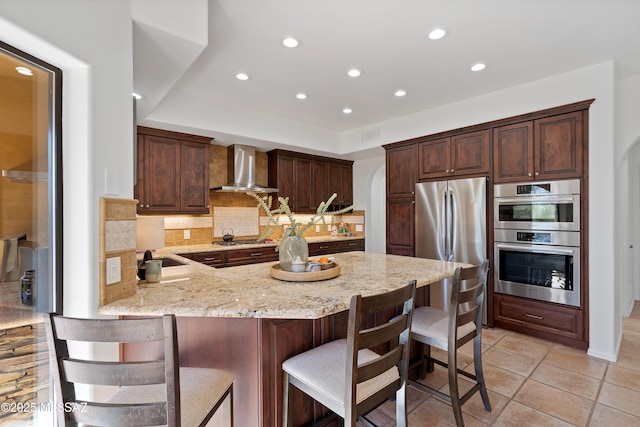 kitchen with stainless steel appliances, wall chimney exhaust hood, light stone countertops, a kitchen bar, and decorative backsplash