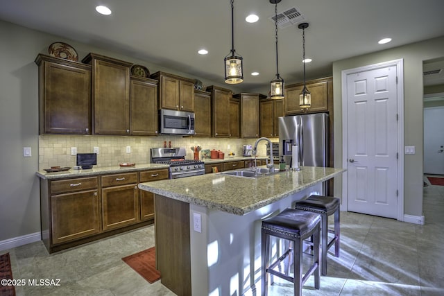 kitchen with pendant lighting, sink, appliances with stainless steel finishes, a kitchen island with sink, and light stone counters