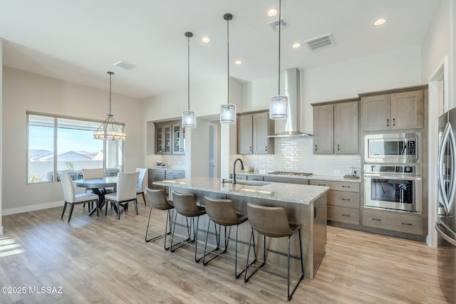 kitchen featuring appliances with stainless steel finishes, pendant lighting, sink, wall chimney range hood, and a center island with sink