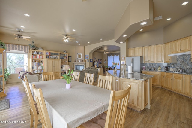 dining space featuring a glass covered fireplace, arched walkways, light wood finished floors, and ceiling fan