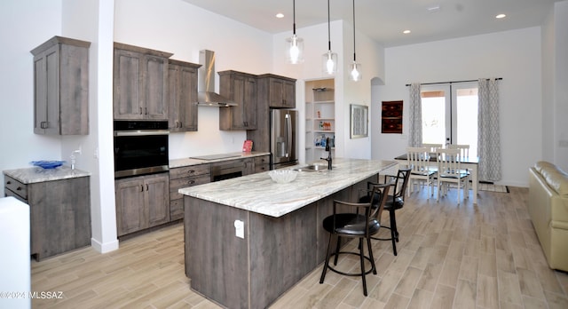 kitchen with pendant lighting, wall chimney range hood, sink, a kitchen island with sink, and stainless steel appliances