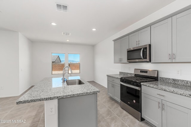 kitchen with gray cabinets, appliances with stainless steel finishes, sink, a kitchen island with sink, and light stone counters